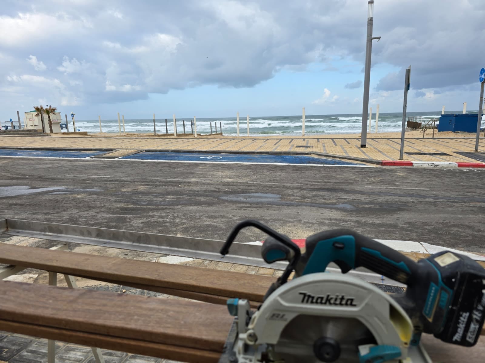 לוחות עץ ארוכים ומסור עגול על חמורי עבודה בצד טיילת, עם חוף וים גלי ברקע