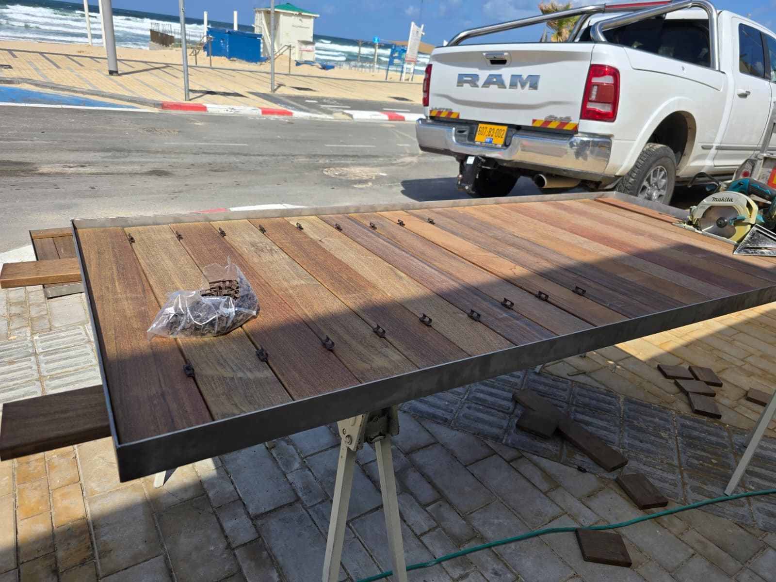 מסגרת נירוסטה שטוחה על חמורי עבודה ובתוכה לוחות איפאה, עם שקית מהדקים על הלוחות וטנדר לבן ברקע