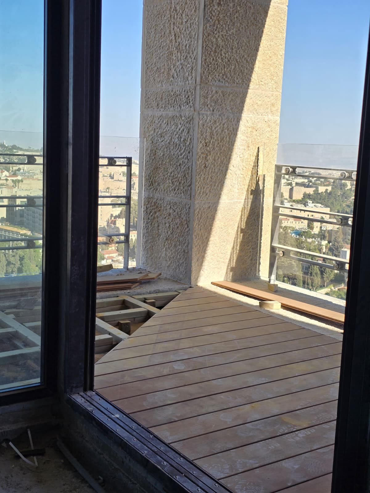 מבט מבעד לדלת הזזה אל דק עץ כהה שהונח באלכסון במרפסת פנטהאוז, לצד עמוד אבן ירושלמית ומעקה זכוכית