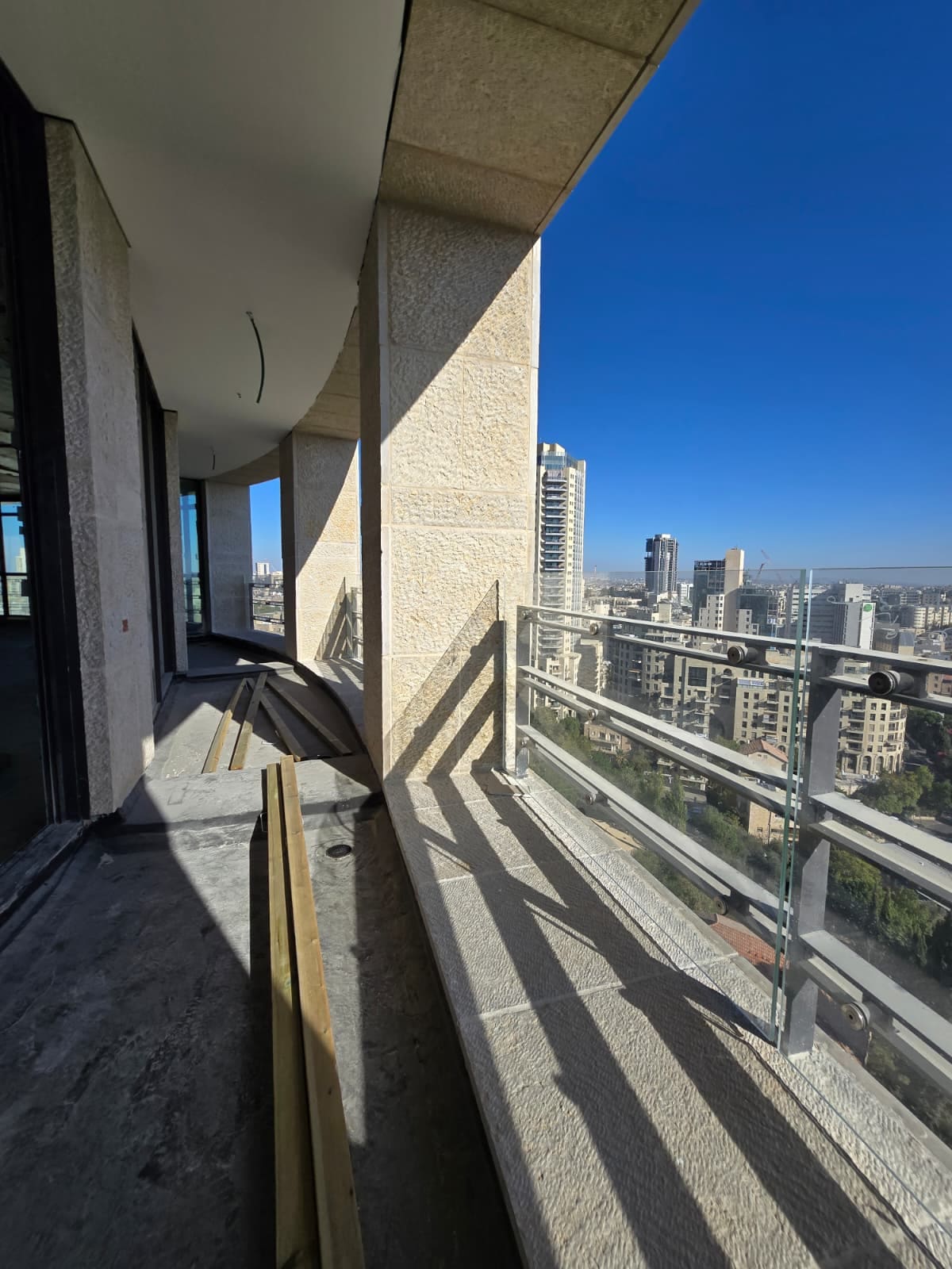 מרפסת פנטהאוז מעוקלת עם רצפת בטון חשופה, עמודי אבן ירושלמית ומעקה זכוכית מול נוף העיר