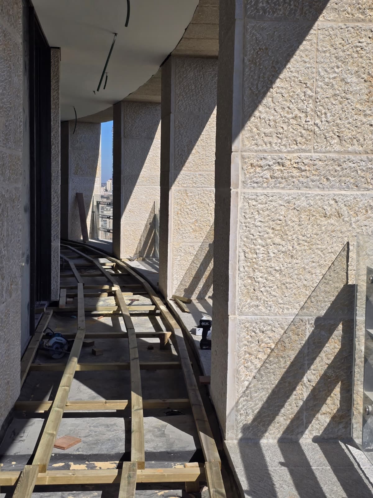 קורת עץ היקפית מעוקלת לאורך מרפסת צרה וארוכה, עם צללי מעקה על קיר אבן ירושלמית