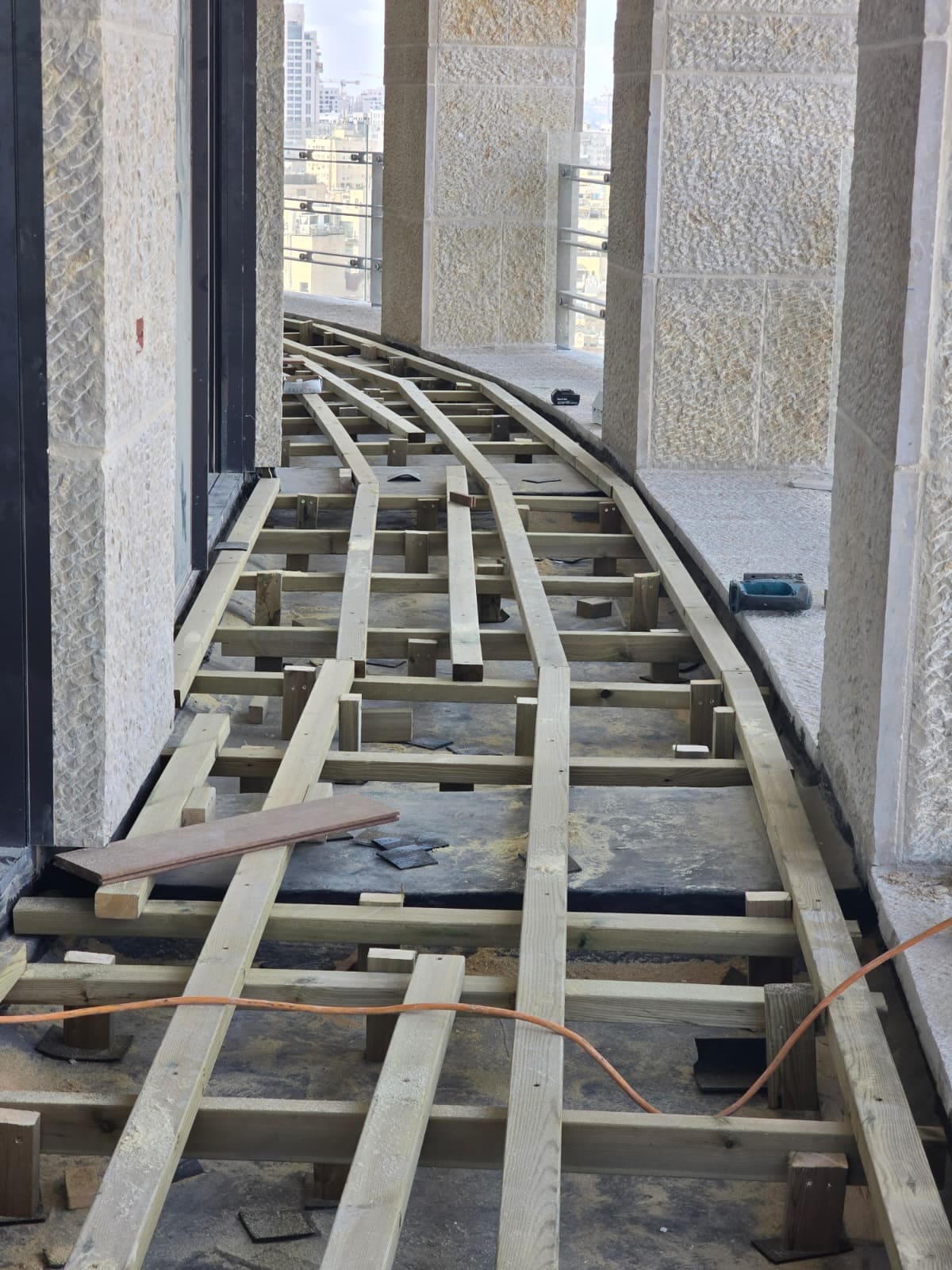 שלד קורות עץ מוגבהות על סדים מעל רצפת איטום, מתעקל לאורך מרפסת בין עמודי אבן