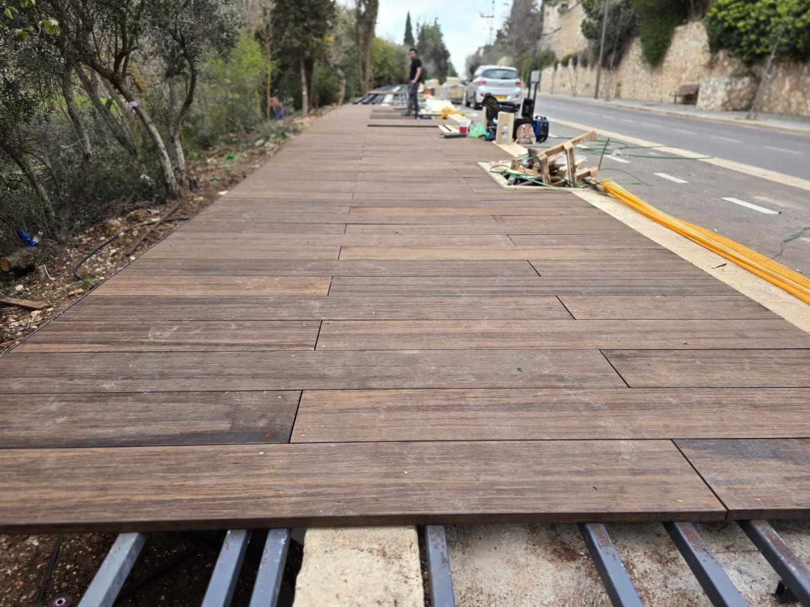 טיילת עץ רחבה לאורך כביש, עם קורות פלדה חשופות בקדמת התמונה וצמחייה משמאל